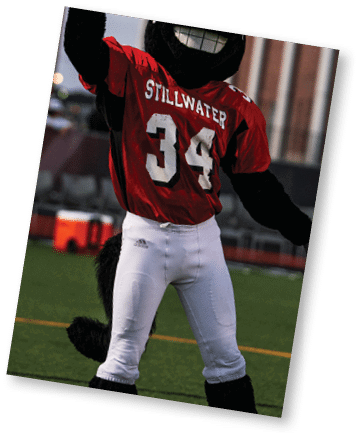 A person wearing a black and red outfit is raising one hand with a finger pointing up on a sports field with a brick archway and bleachers in the background. Description generated by AI