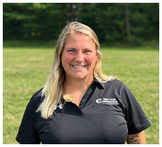 A person with long light hair is smiling and wearing a black polo shirt with a logo that reads Twin Cities Orthopedics Sports Medicine, standing outdoors with grass and trees in the background. Description generated by AI