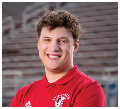 A person with short curly hair is smiling and wearing a red collared shirt against a blurred background. Description generated by AI