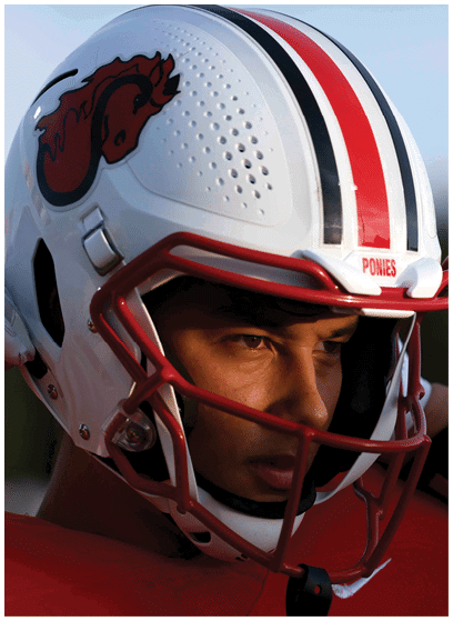 A person wearing a white football helmet with a red faceguard and a red jersey is adjusting the helmet with a gloved hand. Description generated by AI