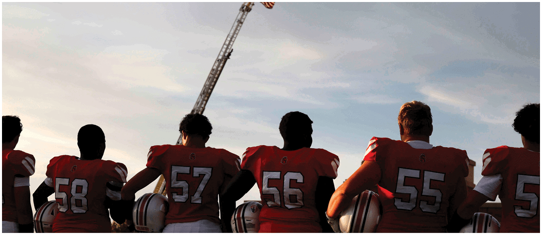 Four people wearing red football jerseys with numbers 57, 56, and 55 are standing with their backs to the camera, holding helmets, while an American flag is raised on a fire truck ladder in the background against a cloudy sky. Description generated by AI