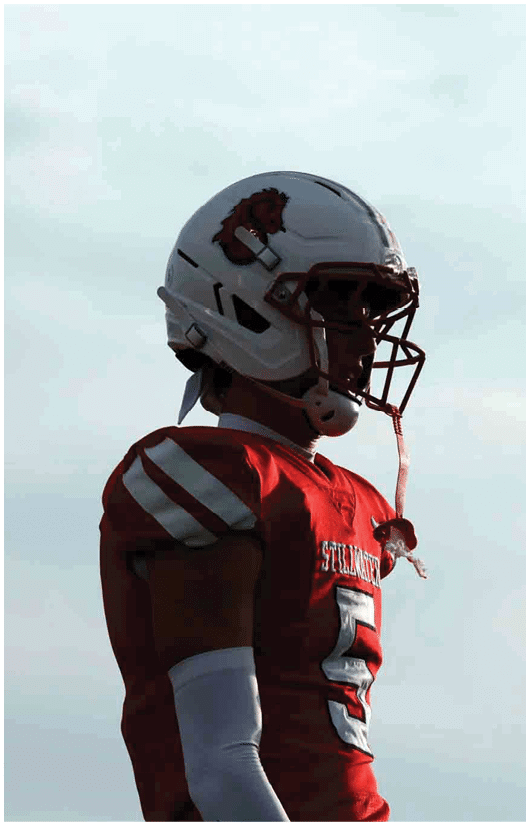 Two people in red football uniforms and helmets stand on a field with empty bleachers in the background. Description generated by AI