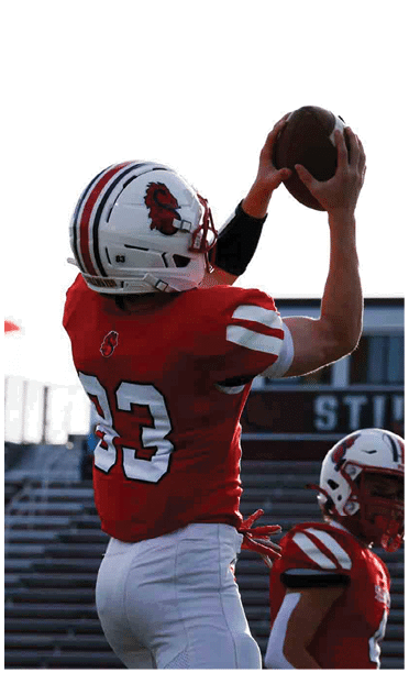A person wearing a red football jersey with the number 33 and a white helmet is reaching up, likely to catch a ball, with empty bleachers in the background. Description generated by AI