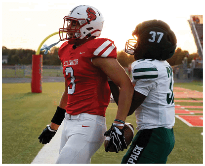 A person wearing a red football jersey with the number 3 and white pants is holding a football while standing on a football field near the end zone. Description generated by AI