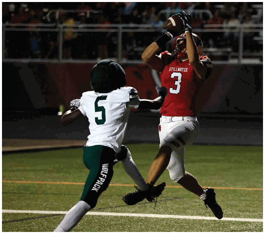 A person in a red football uniform is jumping to catch a football while another person in a white and green uniform attempts to defend on a lit football field at night. Description generated by AI