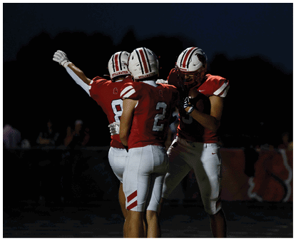 Three people wearing red and white football uniforms are standing together on a field at night, with one person raising an arm. Description generated by AI