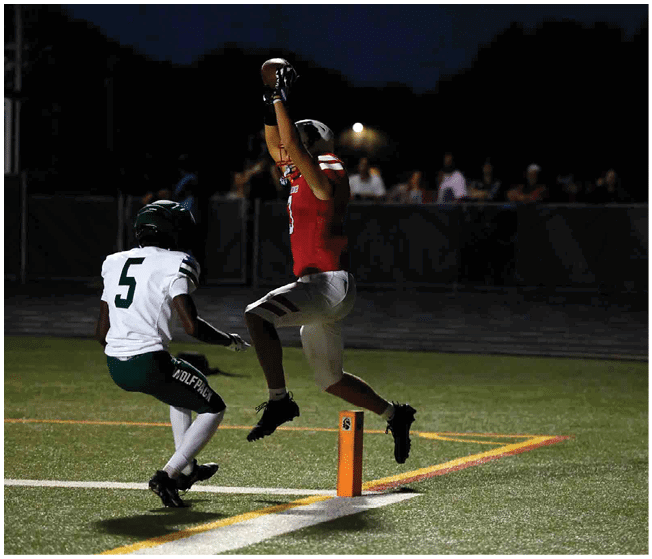 A person in a red sports uniform jumps to catch a football near the end zone while another person in a white and green uniform approaches on a lit football field at night. Description generated by AI