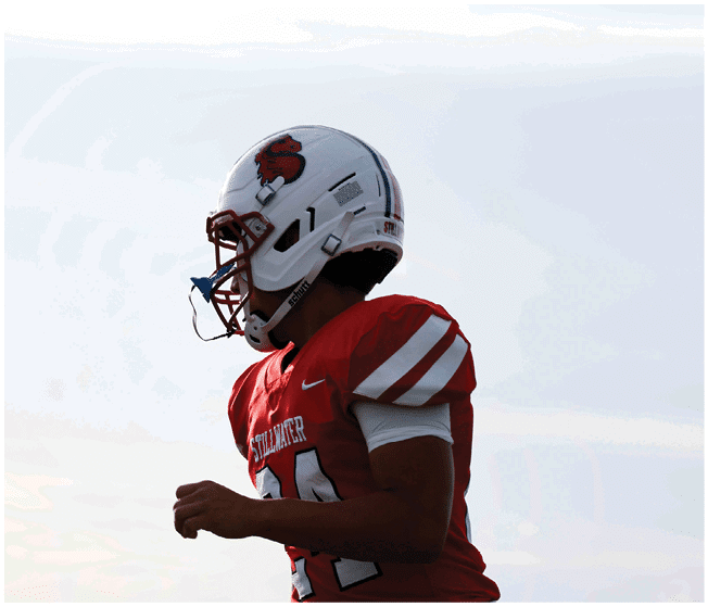A person wearing a red football jersey and white helmet with a blue mouthguard is standing outdoors against a bright sky. Description generated by AI