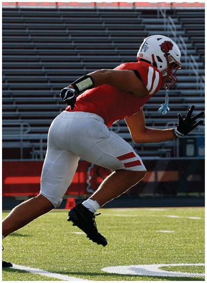 A person wearing a red sports jersey, white pants, and a white helmet is running on a football field with empty bleachers in the background. Description generated by AI