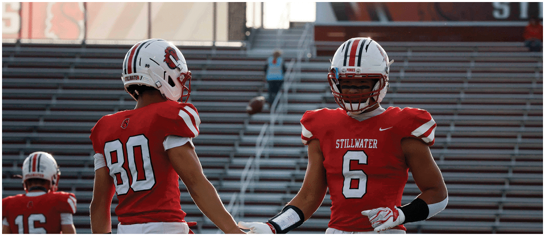 Two adults wearing red and white football uniforms are reaching out to touch hands on a football field with empty bleachers in the background. Description generated by AI