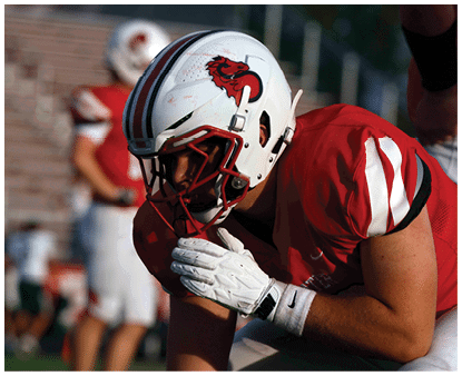 A person wearing a white football helmet and red jersey is crouched in a ready position on a football field with other players blurred in the background. Description generated by AI