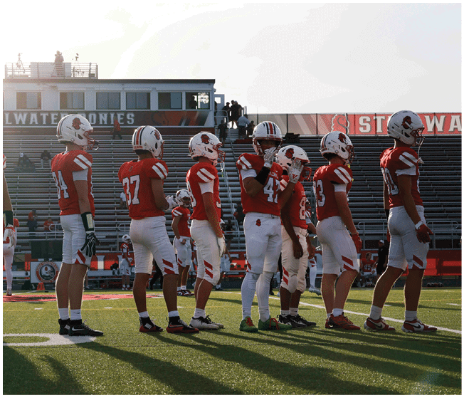 A group of people wearing red and white football uniforms stand in a line on a football field with bleachers and a scoreboard in the background. Description generated by AI