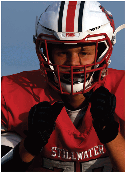 A person wearing a white football helmet with red and black stripes and a red jersey with the word “STILLWATER" is adjusting their black gloves. Description generated by AI