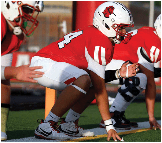 Three people in red and white football uniforms are crouched in a ready position on a green field. Description generated by AI