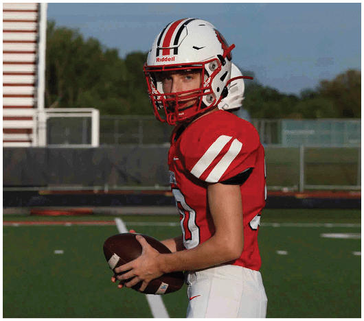 A person wearing a red and white football uniform and helmet is holding a football on a sports field. Description generated by AI