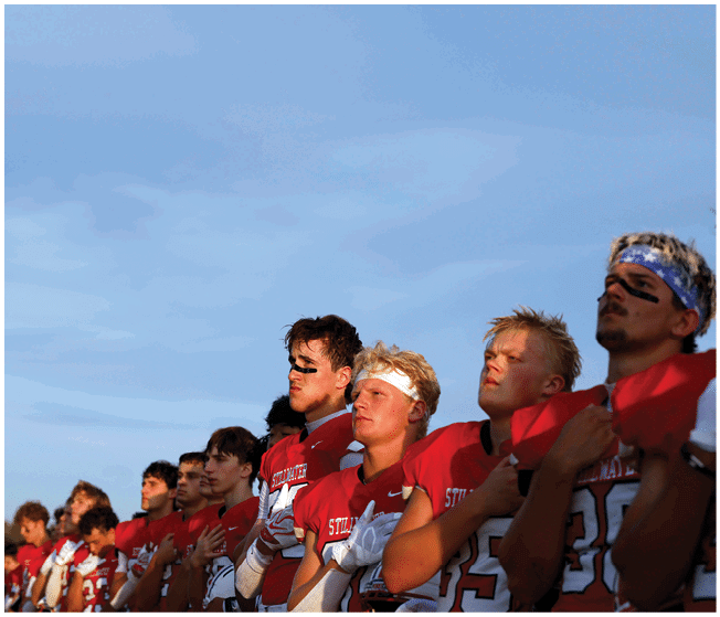 A line of people wearing red sports jerseys stand outdoors with their hands over their hearts against a clear blue sky. Description generated by AI