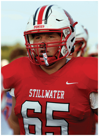 A person wearing a red football jersey with the number 65 and a white helmet labeled “Ponies" is standing outdoors, appearing focused. Description generated by AI