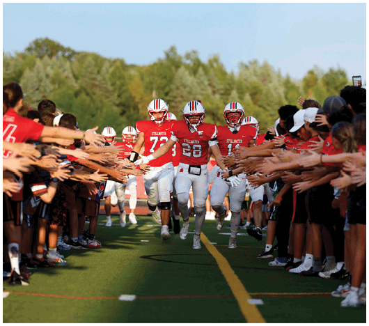 People in red and white football uniforms run down a field between two lines of spectators reaching out to them. Description generated by AI