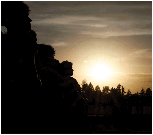 A group of people standing closely together outdoors at sunset with trees silhouetted against the sky. Description generated by AI