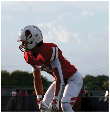 A person in a red and white football uniform and helmet is crouched on a field with other players and trees in the background. Description generated by AI