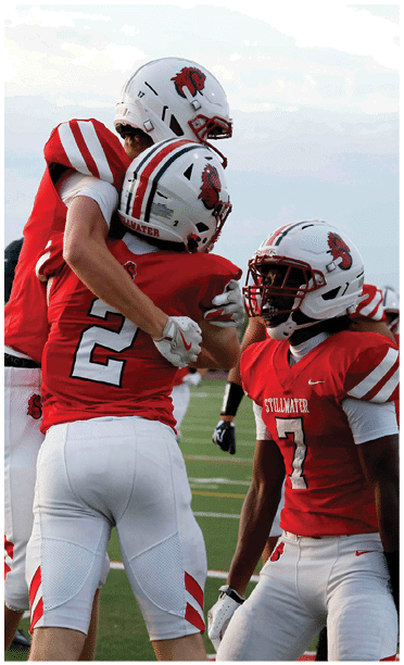 Two football players in red and white uniforms are embracing on a field while players in green and white uniforms stand nearby. Description generated by AI