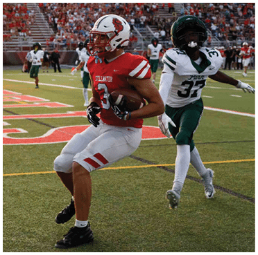 A person in a red football uniform is holding a football and appears to be running or preparing to catch while another person in a white and green uniform is closely pursuing on a football field. Description generated by AI