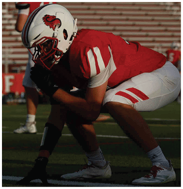 A person in a red and white football uniform is crouched in a ready position on a football field with empty bleachers in the background. Description generated by AI