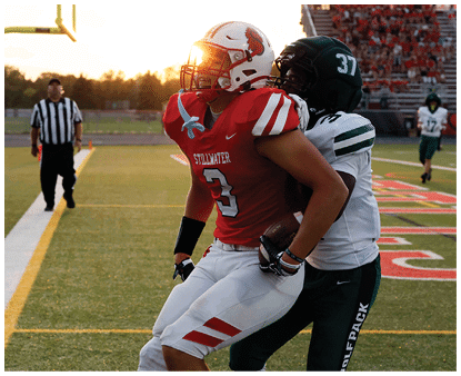 Two people in football uniforms are engaged in a tackle on a field during a game with a referee and spectators in the background. Description generated by AI