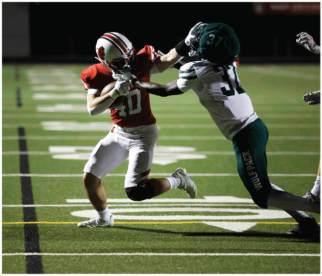 Two people in football uniforms are engaged in a play on a field, with one person in a red jersey holding the ball while the other in a white and green jersey attempts to block or tackle. Description generated by AI