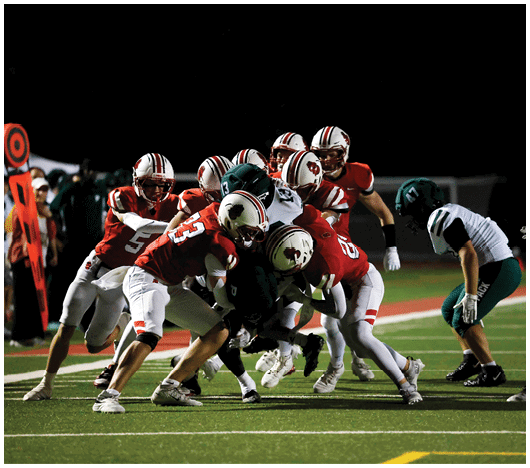 A group of people wearing red and white football uniforms are tackling a person in a dark green uniform on a football field at night. Description generated by AI