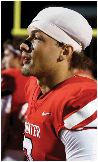 A person wearing a red football jersey with white stripes and a white head covering is standing on a field, looking to the side with black sports tape on their nose. Description generated by AI