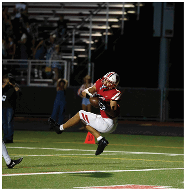A person in a red and white football uniform is airborne while catching a football on a field at night with spectators in the background. Description generated by AI