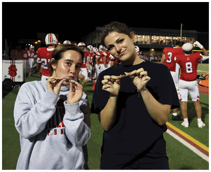 Two people stand on a football field at night holding small objects near their mouths while players in red uniforms and helmets are visible in the background. Description generated by AI