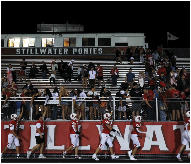 People in sports uniforms walk along a track in front of a crowd seated on bleachers under a sign that reads “Stillwater Ponies" at night. Description generated by AI