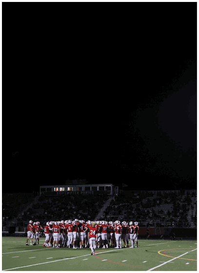 A group of people wearing red and white sports uniforms are gathered on a football field at night with stadium seating and a scoreboard visible in the background. Description generated by AI