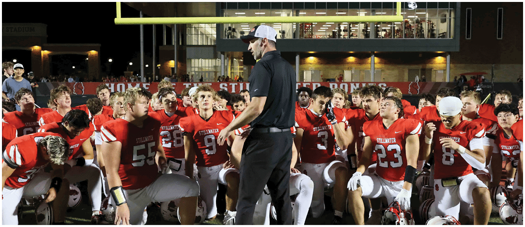 A group of people in red and white football uniforms kneel on a field around a person in black clothing and a white cap who appears to be giving instructions under stadium lights at night. Description generated by AI