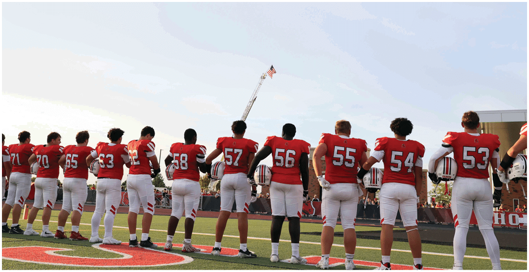 Seven people in red football jerseys and white pants stand side by side on a football field holding helmets with a raised flag in the background. Description generated by AI