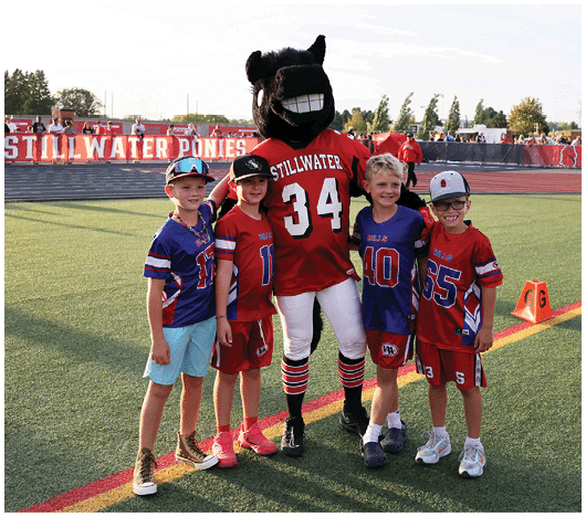 Four children in sports jerseys stand on a football field smiling next to a person in a black horse mascot costume wearing a red jersey with the number 34 and the word Stillwater. Description generated by AI
