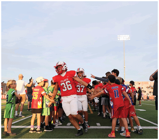 People in sports uniforms and casual clothing are gathered on a football field with some players running through a line of people giving high-fives. Description generated by AI