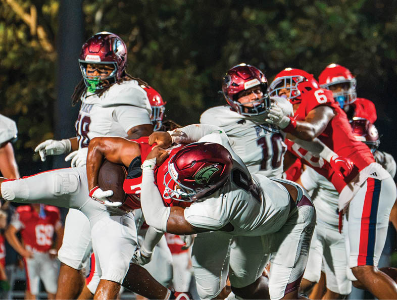 People wearing football helmets and uniforms are engaged in a tackle during a game on a field with a blurred background. Description generated by AI