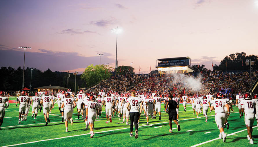 A group of people in white sports uniforms with red helmets are running onto a green football field toward a stadium filled with spectators during a game. Description generated by AI
