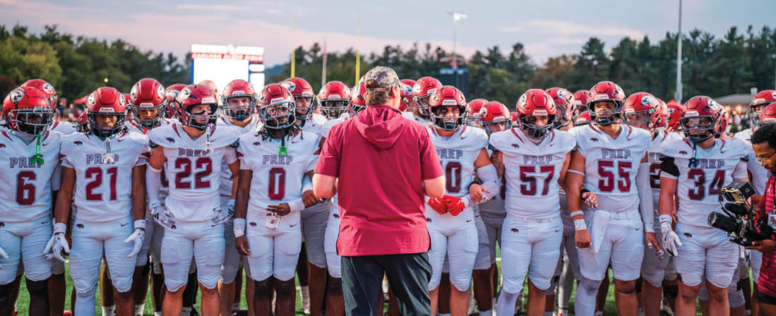 A group of people wearing white football uniforms stand in a line on a field facing a person in a red shirt and dark pants. Description generated by AI