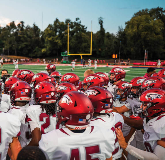 A group of football players wearing red helmets and white jerseys are gathered closely on a field near a goalpost with trees in the background. Description generated by AI