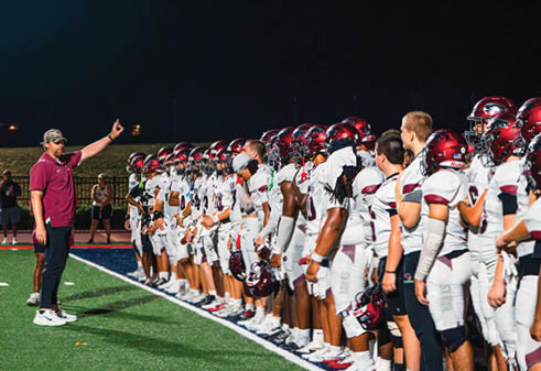 A group of people in white sports uniforms stand in a line on a football field while a person in casual clothing gestures toward them. Description generated by AI