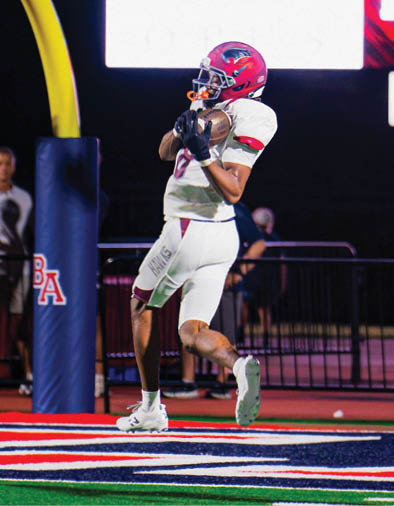 A person in a white football uniform catches a pass near the goalpost while another person in a red football uniform runs toward them on a brightly lit field at night. Description generated by AI