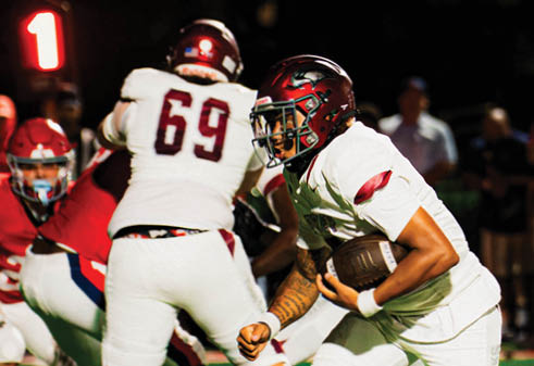 A person in a white football uniform is running with the ball on a green field while other players and people stand in the background. Description generated by AI