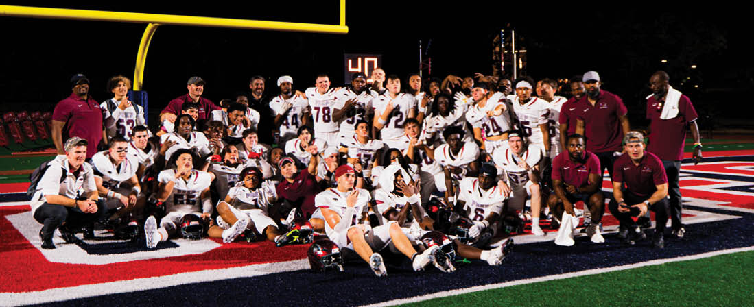 A large group of people wearing sports uniforms and casual clothing are gathered on a football field posing for a team photo. Description generated by AI