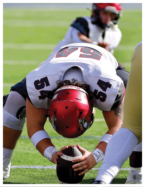 A person wearing a red helmet and white football uniform with the number 54 is crouched on a football field holding a football, preparing to snap it, with another player in white and gold uniform in front. Description generated by AI