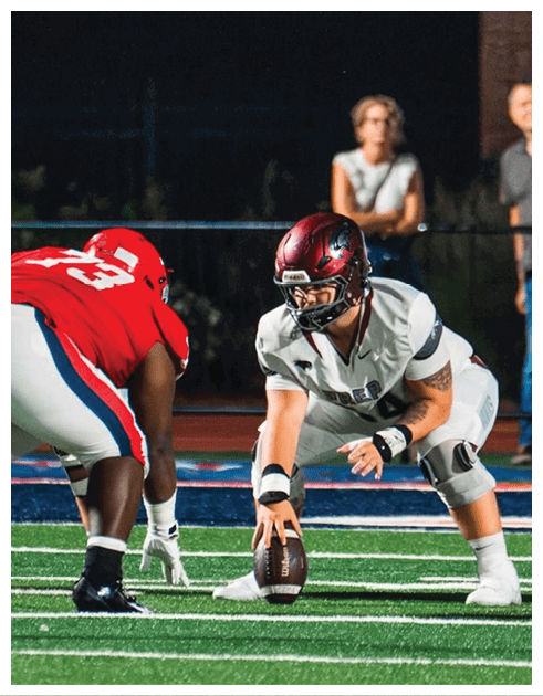 Two athletes in football uniforms face each other on a field with two adults standing in the background. Description generated by AI