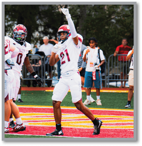 Two people in football uniforms and helmets are on a field, with one raising an arm and pointing upward. Description generated by AI
